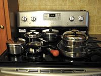 Photo showing seven pieces of cookware on stovetop including various sizes of stainless steel pots and pans with lids.