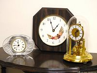 Three clocks displayed on a wooden surface: the Cristal D'Arques quartz crystal desk clock with carved roses on the left, the Tempo octagonal wood wall clock in center, and the vintage German Kern 400 day torsion clock with glass dome on right.