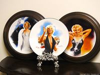 Photo showing all three Marilyn Monroe collectible plates: two are wood-framed and one is unframed on a decorative stand, placed on a dark wooden surface.