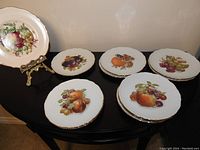 View of ten German Bavaria Schumann Arzberg porcelain dessert plates with gilded edges and fruit designs, displayed on a black table.