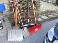 Five shovels leaning against a bush on pavement: two large aluminum bladed snow shovels with wood handles, a red plastic snow shovel with metal handle, and two garden shovels with wooden handles.