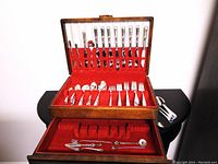 Photo of the opened wooden storage chest showing multiple rows of knives in the lid, forks, spoons, and extra forks in drawer with red lining.