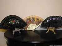 Three ladies' fans displayed on a black surface, including two black hand-painted fans and one vintage fabric and celluloid fan with lace and painted scene