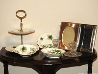 Overall view of all items on a dark wood table showing two-tiered cake tray, three bone china dishes with green trillium floral pattern, crystal candle holders, and two metal photo frames.