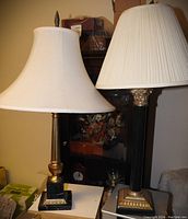 Pair of stone column table lamps with classical Corinthian and reed motifs, black marble bases with gilt accents, located on furniture surfaces indoors.