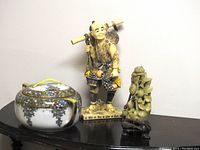 Wide view of all three items on a wooden surface showing the farmer figure, jade-like carving, and covered dish together.