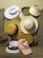 Nine women's hats of assorted styles laid out on a fabric surface: white sports hats, straw hats, visors, a pink baseball cap, and cloth hats.