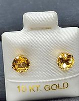 Close-up front view of the pair of 10k yellow gold stud earrings with round faceted citrine stones mounted in 10k gold backs held in a white display holder with '10 KT. GOLD' label.