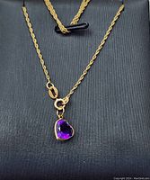 Photo of a delicate gold chain necklace presenting the heart-shaped purple amethyst pendant on black background.