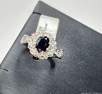 Close-up top angle view of 925 sterling silver ring with oval blue sapphire surrounded by white topaz on a black display box, showing details of band sides.