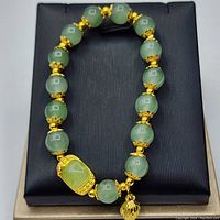 Top-down view of gold plated stainless steel bracelet with smooth green jade beads and central rectangular jade piece, displayed on a black cushion.