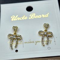 Front view of pair of gold plated stainless steel earrings with heart-shaped stud and bow-shaped dangle design, set with clear cubic zirconia stones, displayed on white card with brand Uncle Beard in gold lettering.