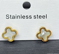 Close up front view of the pair of gold plated stainless steel four leaf clover earrings with mother of pearl inlay displayed on white card labeled 'Stainless steel'.