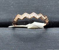 Rose gold stainless steel ring displayed on a black surface, showing the zigzag front design with cubic zirconia stones embedded.