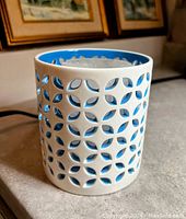 White ceramic candle holder with blue interior and geometric cutouts, displayed on a surface with framed artwork in the background.