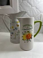 Ceramic pitchers side by side showing floral design on taller pitcher and citrus design with text on shorter pitcher