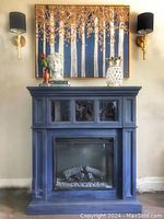 Front view of electric fireplace painted blue with decorative items on mantel, including a head bust, pineapple statue, and books. Shows overall condition and design of fireplace.