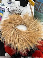 Photo showing a full circular fall wreath made up of fluffy dried wheat and grass stalks on a table, surrounded by miscellaneous items.