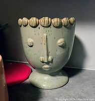 Front view of a beige ceramic flower pot shaped like a human face, with sculpted eyes, nose, lips, and crown-like rim.