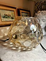 Photo of the brownish glass vase showing its rounded shape and multiple raised bubble patterns on the glass surface. Vase is placed on a table with framed pictures and a decorative bust in the background.