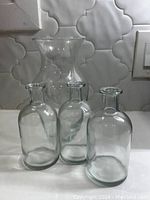 Four clear glass vases displayed on a white surface against a tiled wall, showing different shapes and sizes.