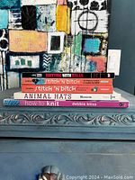 Side view of five knitting books stacked on a table with abstract painting in background.