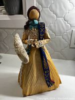 Photo showing the front of the Mexican corn husk doll wearing a yellow lace-trimmed dress, blue sash, and holding a lace-decorated straw hat.