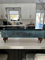 Full view of teal chalk painted rectangular wooden box on kitchen countertop, showing carved decorative design and four wooden legs.