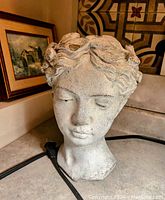 Concrete plant vase in the shape of a human head with detailed features and wavy hair, gray color with weathered texture.