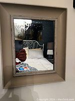 Photo of small wood framed mirror hanging on wall with reflection of bed and lamp.