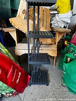 Small, black, wooden stand with four slatted shelves shown in front of a wooden chair and cluttered background.