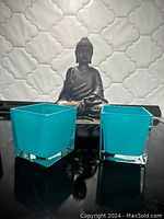 Two blue glass square tea candle holders with clear bases on a reflective surface.