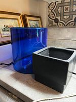 Two plant containers placed side by side on a countertop, one blue glass cylindrical and one black ceramic square shape.