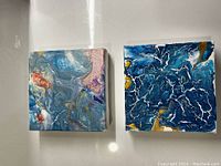 Photo of two original acrylic pour paintings on small square canvases, placed side by side on a white surface.