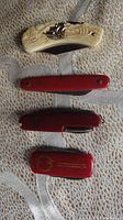 Four assorted pocket tools displayed closed, showing ivory-style handle with a ship, red slim knife, red multi-tool, and red advertising tool.
