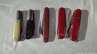 Five pocket knives lying flat, showing red Swiss Army style knives and two traditional Barlow style knives in cream and black handles.