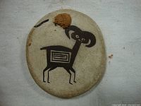 Front of the round art disc showing black geometric ram painting on beige surface with worn patches.