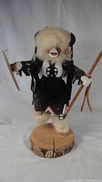 Front view of the Kachina Buffalo Warrior figurine showing detailed face, fur headdress, costume, sticks, and shield on a wooden base.