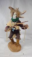 Front view of Kachina doll showing painted mask, green collar, feathers, bow and arrow, and base