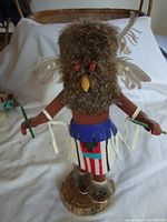 Front view of the Kachina doll showing fur-covered owl head, feather wings, fringed belt, decorated skirt, and green sticks held in hands.