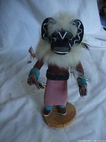 Front view of the Uncle Black Ogre Kachina doll showing detailed painted face, white fur headdress, pink skirt, turquoise wrist and ankle cuffs, feathers on wrists, standing on a wooden base.