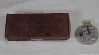 Brown leather rectangular case with visible wear and 'W.G. Neill & Co. Ltd' embossed on top, next to a small oval Chinese medicine bottle with colorful painted designs and stopper.