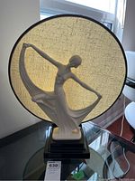 Full view of the lamp showing the dancing female figure in glazed white ceramic, black base, and large round woven fabric shade in beige with black trim.