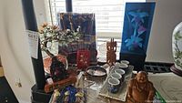 Photo showing two woven mats with chopstick and soy sauce holders, porcelain sake set including multiple cups on a tray, wooden carved figures including Buddha and another statue, and fiber optic floral display in blue case