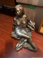 Front view of the painted sculpture showing seated female figure on rocky base, details of face, hair, dress, and pose.