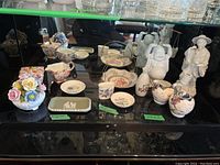 Front and top overview photo showing the ceramic figurines, trinket trays, dishes, cream and sugar sets, and a variety of floral-decorated ceramic pieces arranged on a glass shelf.
