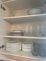 Shelf with stacked white Corelle dinner plates, side plates, clear glass tumblers, and mugs with floral etching. One large white serving bowl on top shelf.