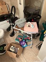 Photo showing black rollator walker with seat and mesh basket, box of health supplies on floor, bedside commode with padded seat, gray bedpan and hygiene products on top.