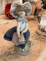 Front view of Lladro figurine depicting a young girl with a large white hat, light blue capelet, blue skirt, and basket of flowers.