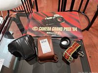 Top view of Nikon Camera Grand Prix '84 placemats, black Nikon leather camera case, brown protective case, lens shade, and colorful woven strap laid out on glass table.